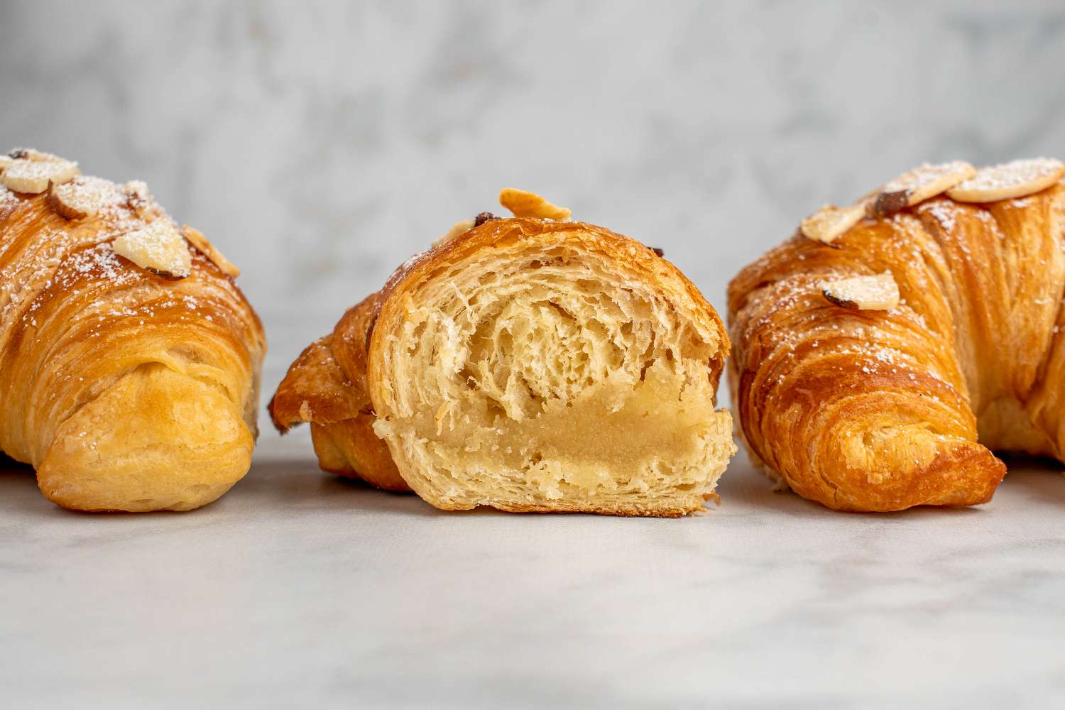 Freshly baked almond croissants cooling on a metal rack on a kitchen counter with a small sifter of powdered sugar nearby