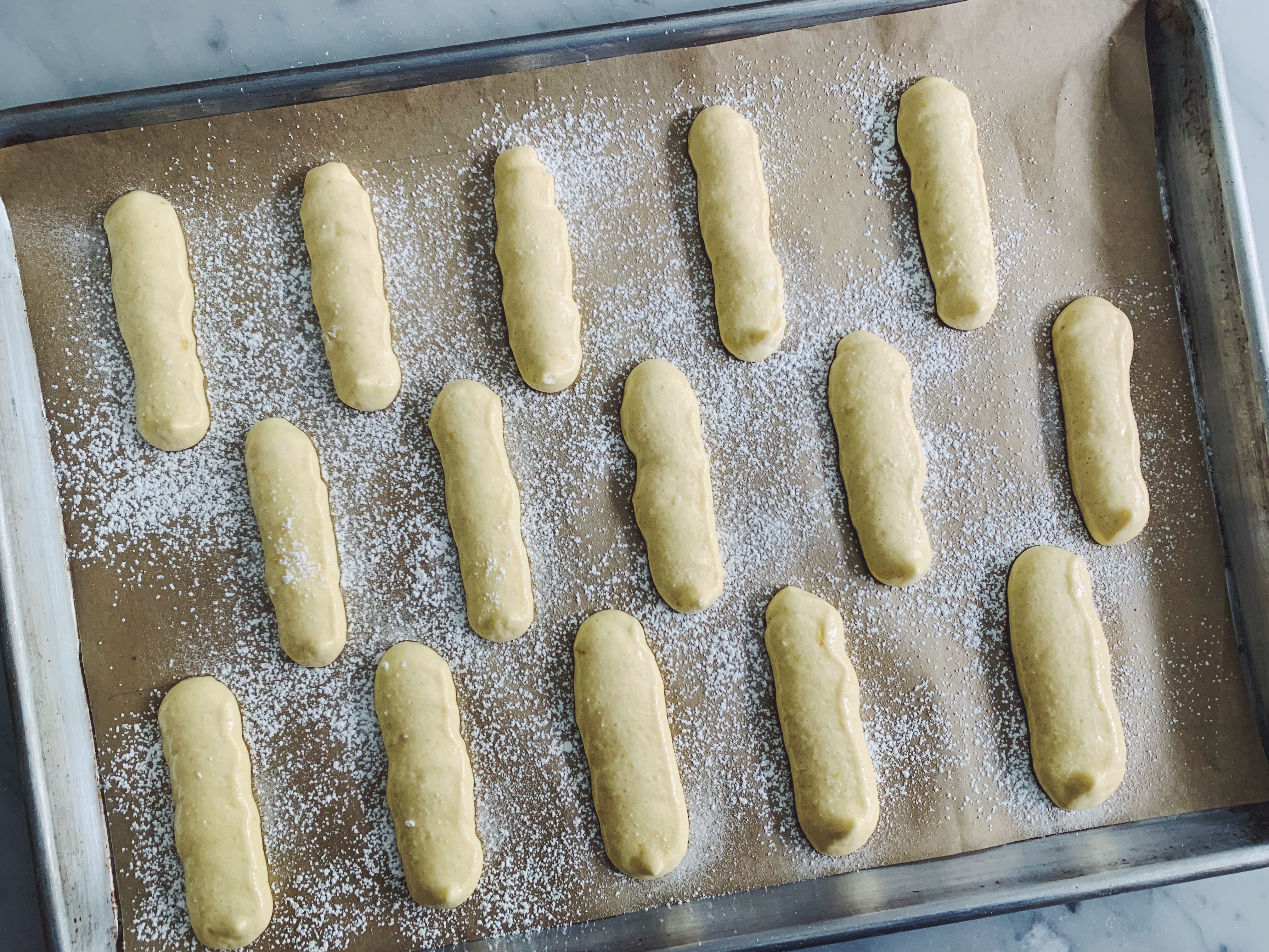 Freshly baked ladyfingers arranged in neat rows on a parchment-lined baking sheet on a warm kitchen counter, golden tops dusted with powdered sugar