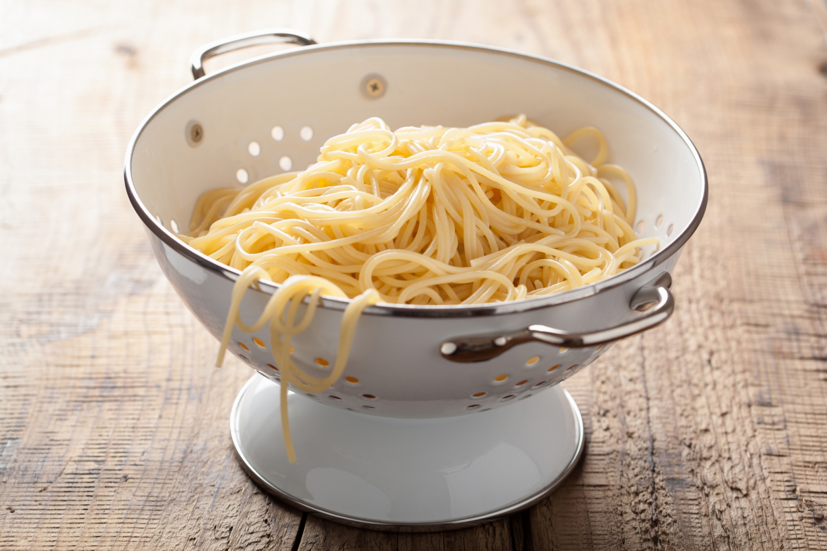 Freshly cooked spaghetti being drained in a metal colander over a sink with steam rising