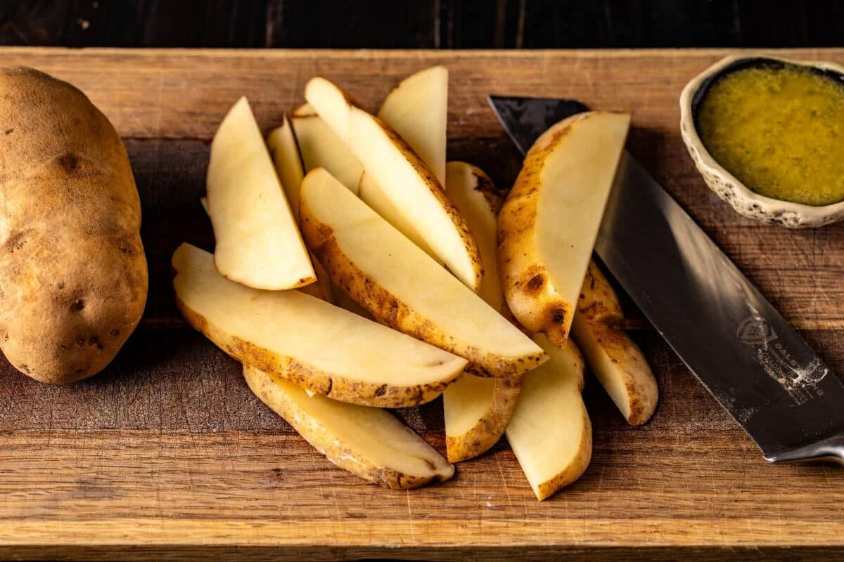 Freshly cut potato wedges spread across a wooden cutting board with a chef's knife nearby on a light kitchen counter