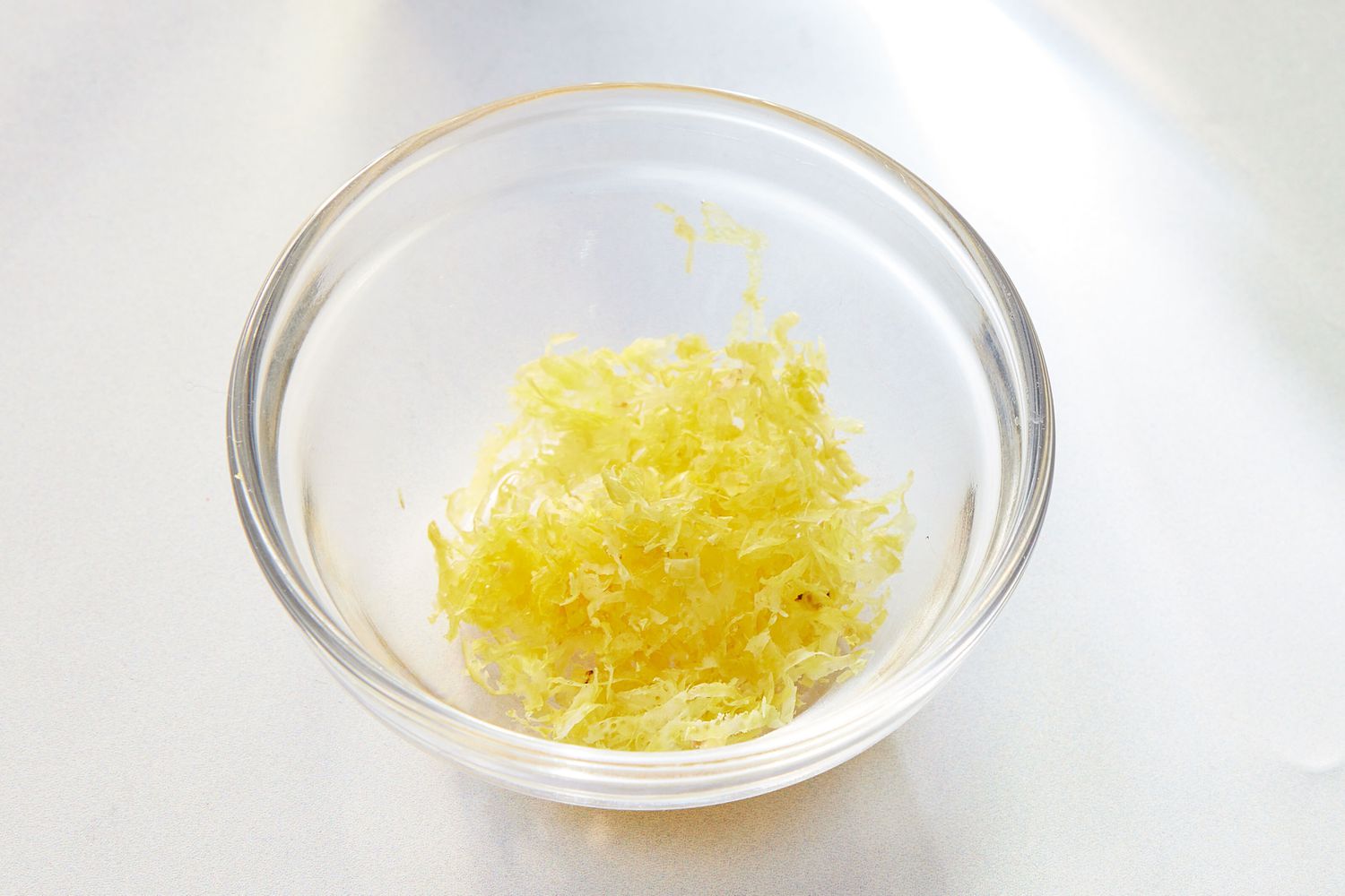 Freshly grated lemon zest in a small bowl next to whole lemons on a wooden cutting board on a kitchen counter