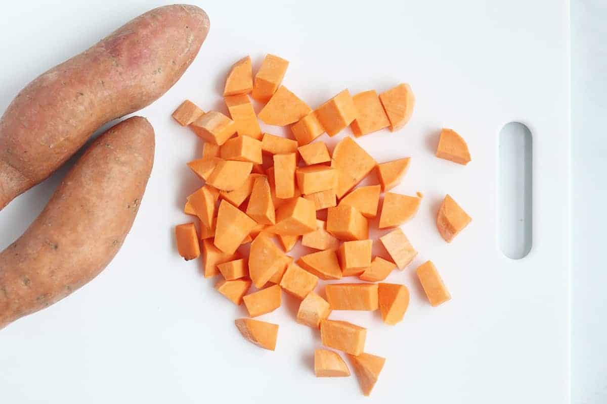 Freshly peeled sweet potatoes cut into small cubes on a wooden cutting board with a chef’s knife nearby