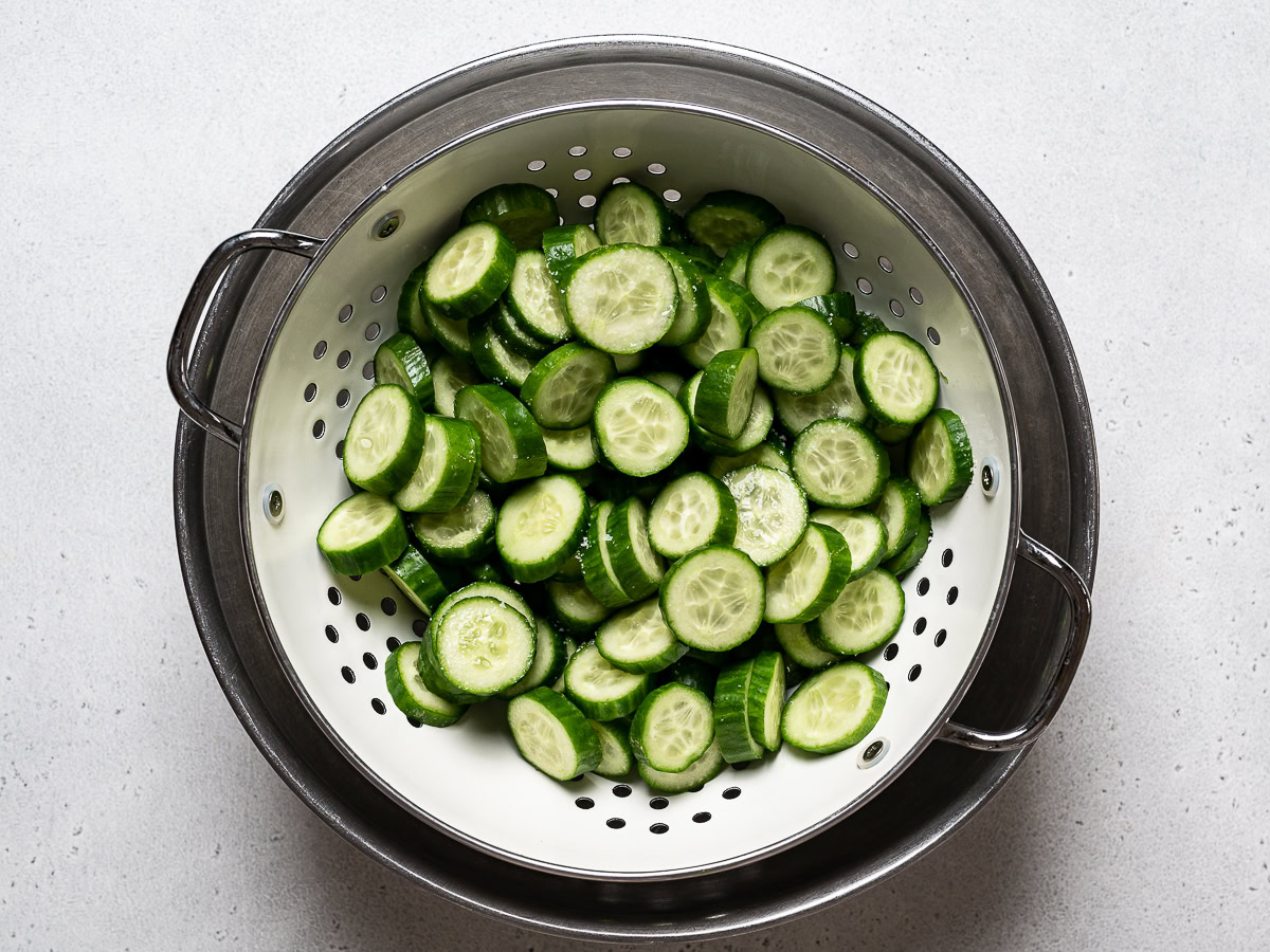 Freshly sliced Persian cucumbers sitting in a colander over a bowl, lightly sprinkled with salt on a kitchen counter