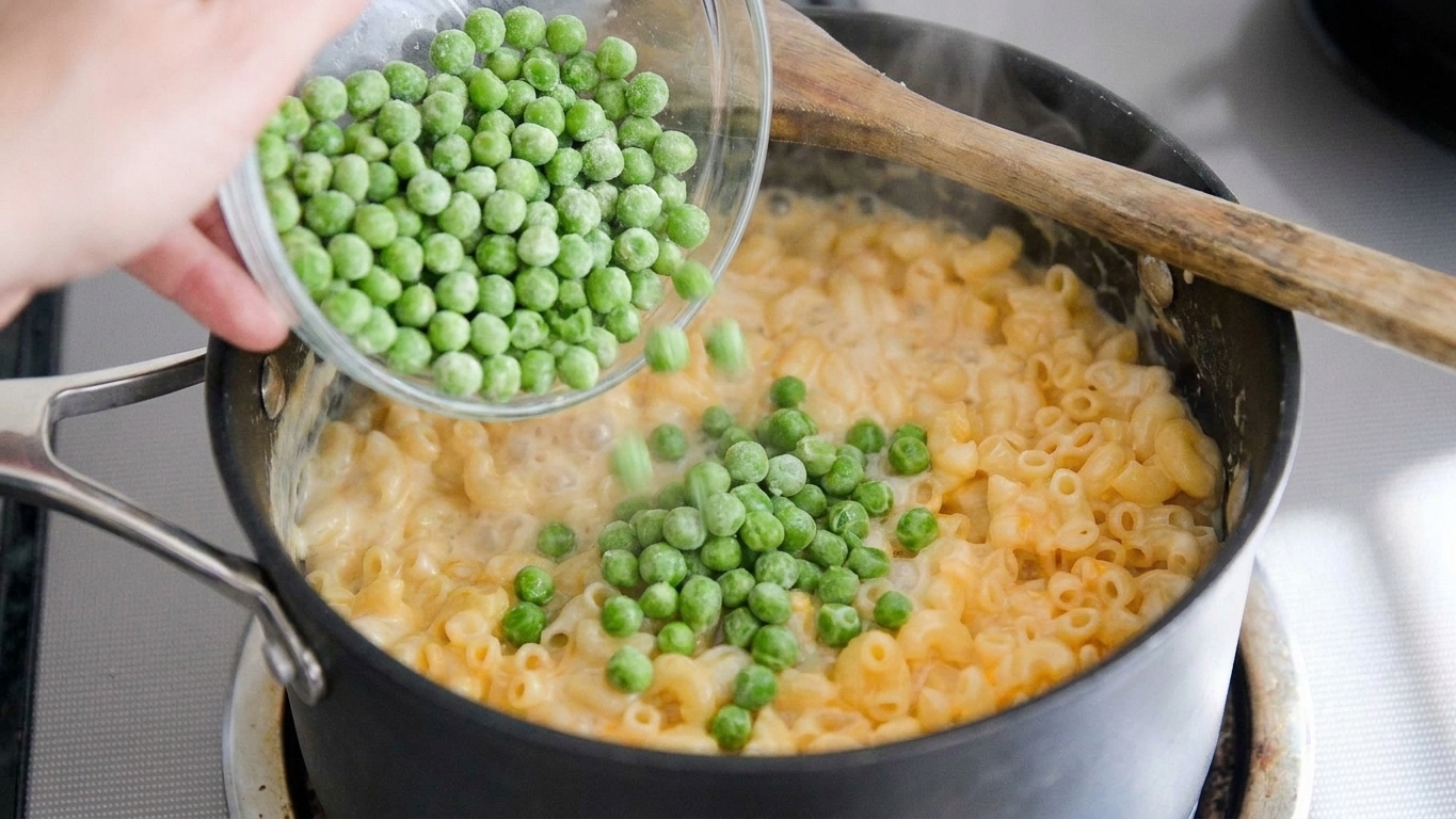 Frozen green peas being poured into a pot of simmering macaroni on the stovetop