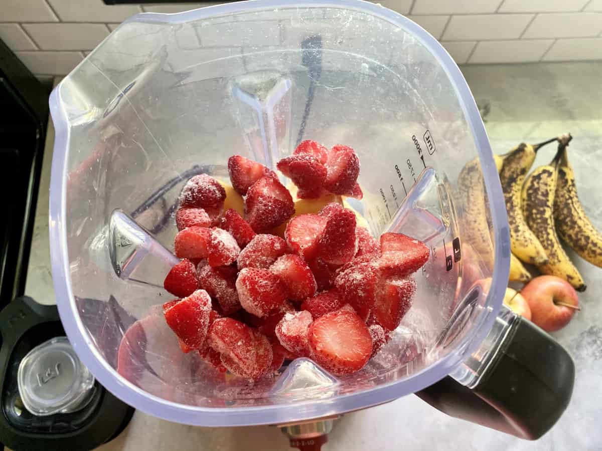 Frozen strawberries and ice measured and added to a blender jar on a clean kitchen counter with a lime and measuring spoons nearby