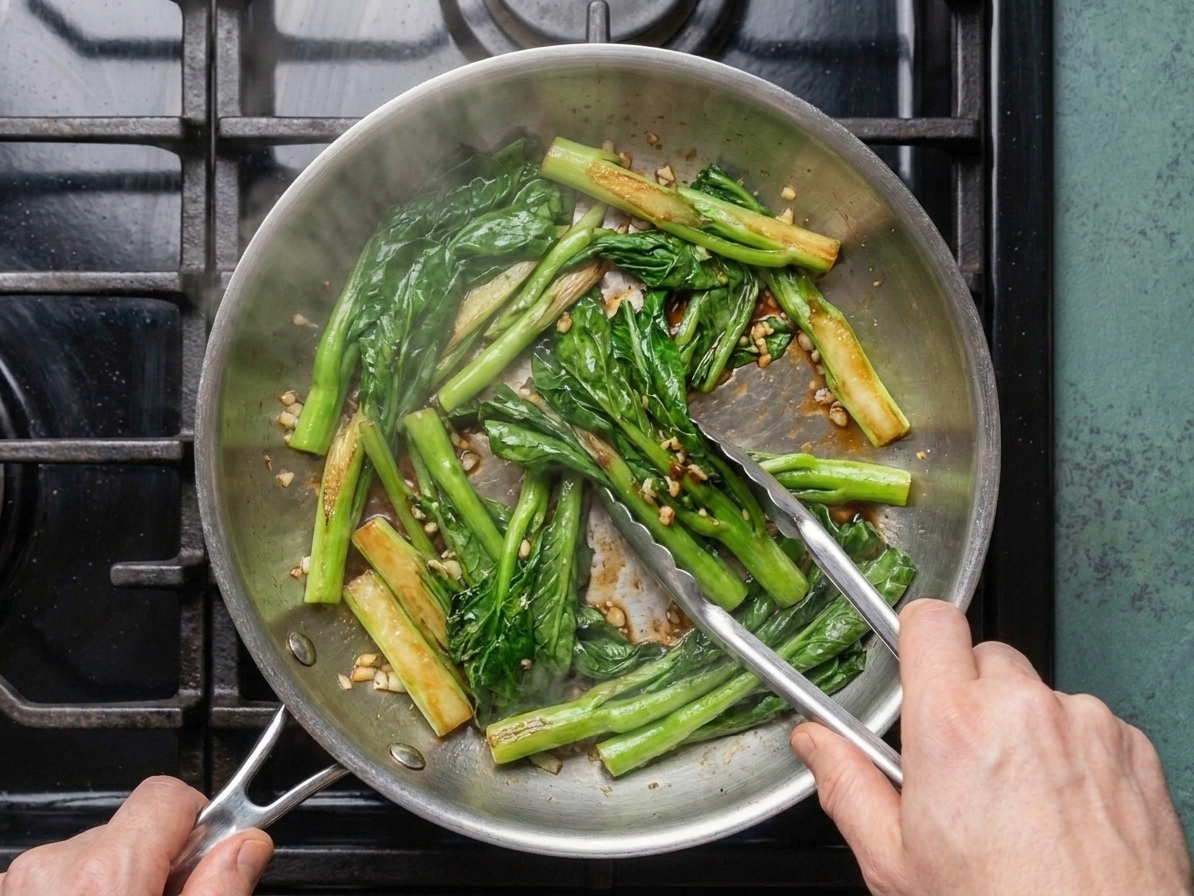 Gai lan being stir-fried in a hot skillet with tongs, showing lightly browned edges on the stems