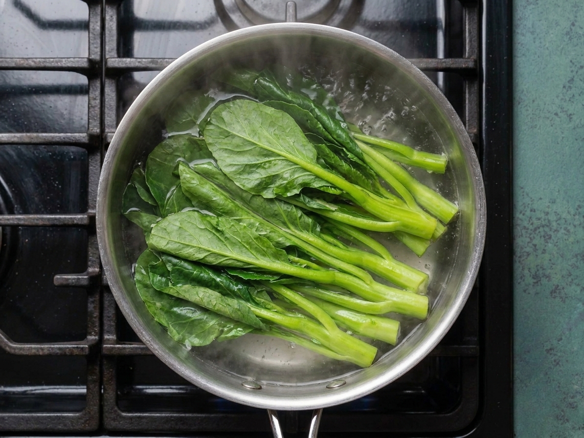 Gai lan stems and leaves blanching in a pot of boiling water on a stovetop with steam rising