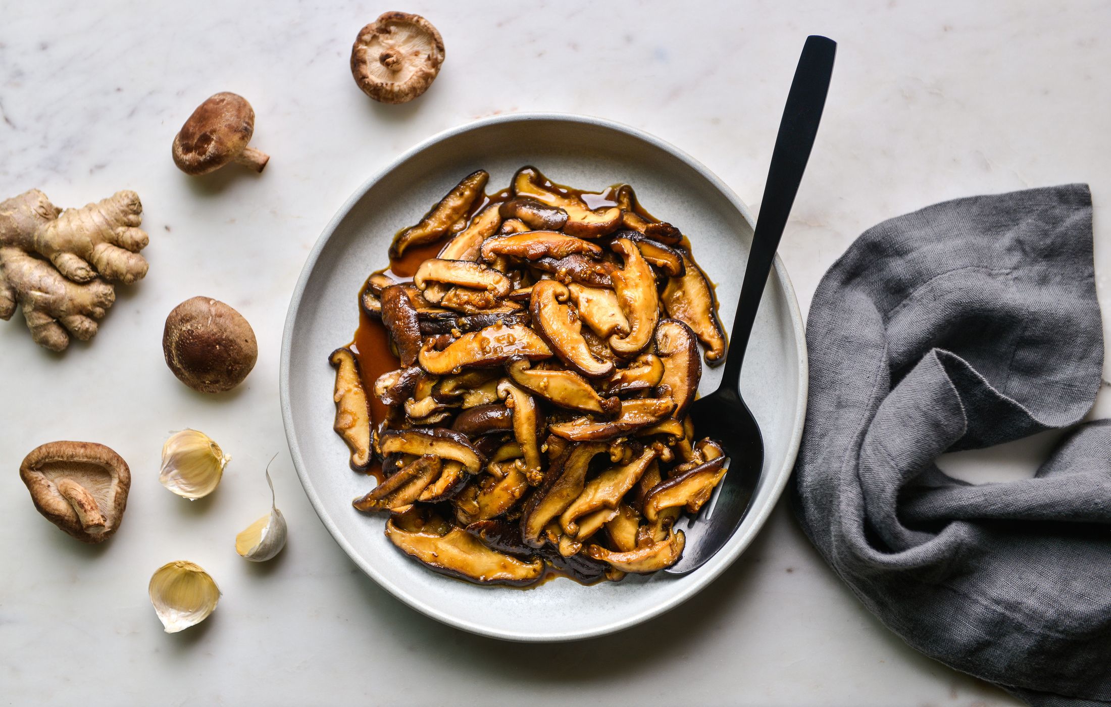 Garlic, grated ginger, and sliced mushrooms sizzling in a pot with a wooden spoon stirring on a stovetop