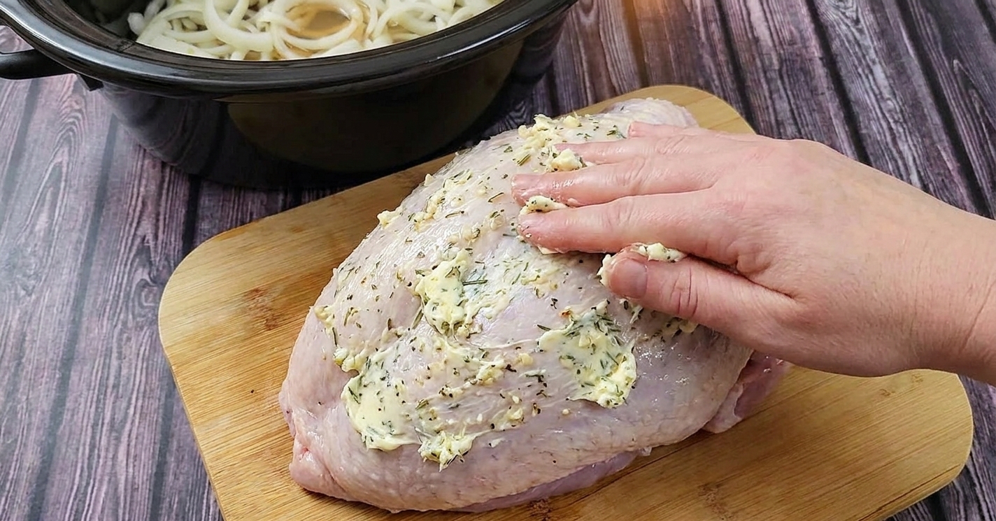 Garlic herb butter being rubbed over a raw turkey breast on a cutting board next to a slow cooker