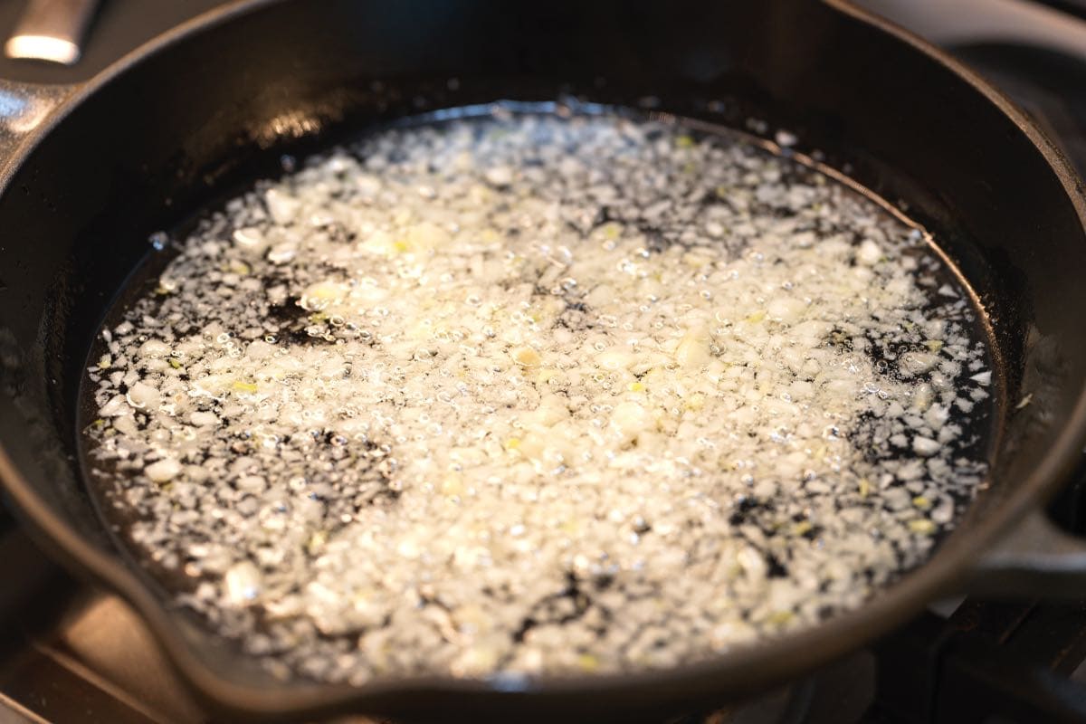 Garlic slices gently frying in a shallow layer of oil in a skillet on a stovetop, with a wooden spoon stirring