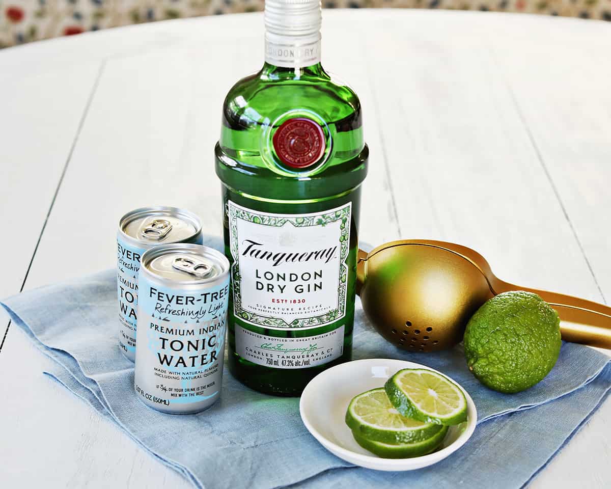 Gin bottle, tonic water bottle, fresh limes, and a jigger arranged on a tidy kitchen counter next to a filled ice tray