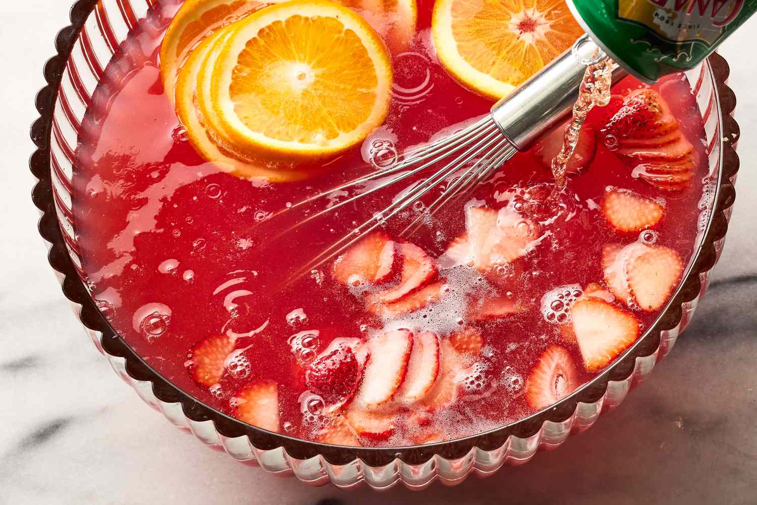 Ginger ale being poured into a large glass punch bowl of red punch with fruit and ice on a kitchen counter