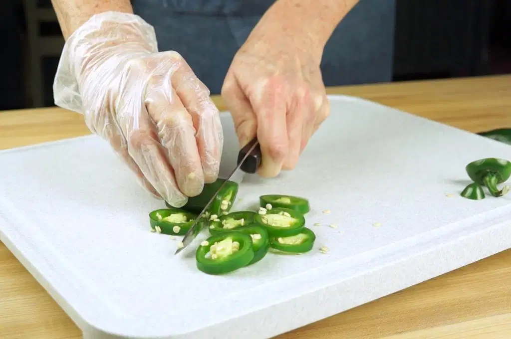 Gloved hands slicing jalapeños into thin rings on a cutting board on a bright kitchen counter