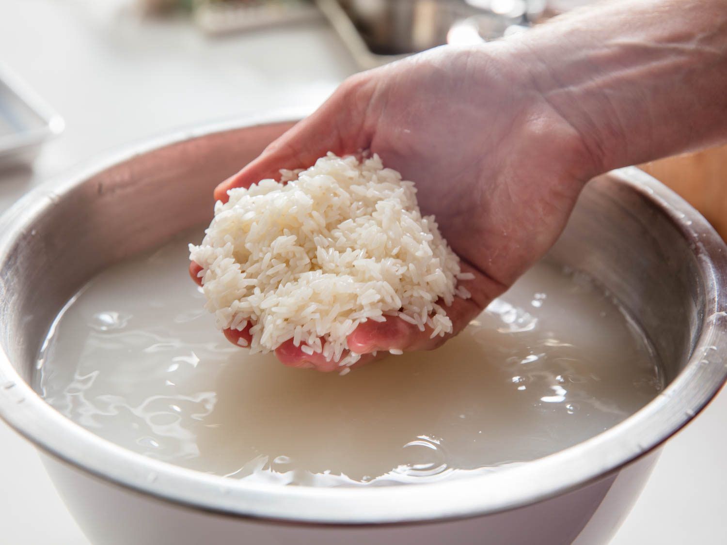 Glutinous rice soaking in a glass bowl of water on a kitchen counter next to a fine mesh strainer