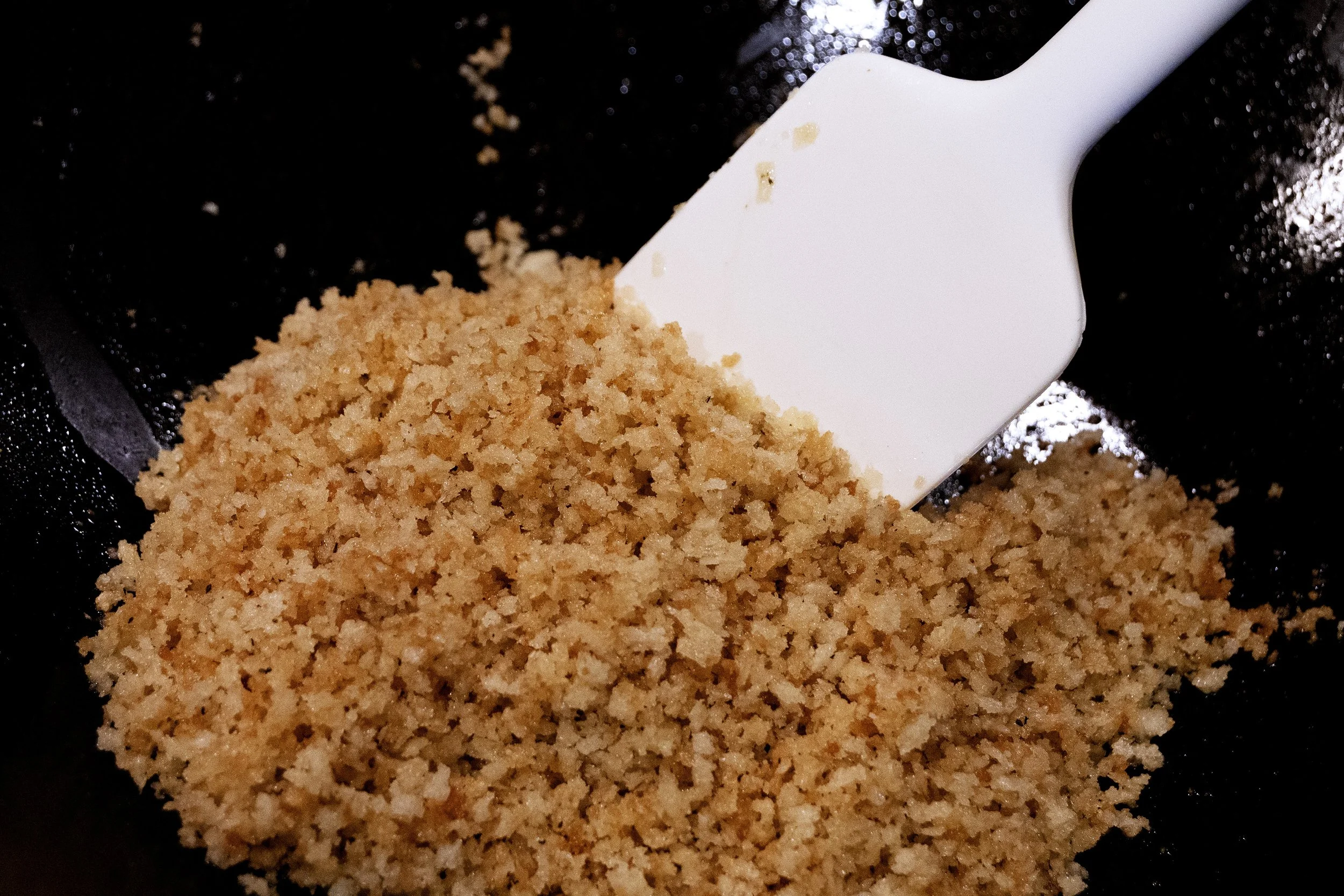 Golden breadcrumbs toasting in a skillet with a wooden spoon stirring on a stovetop