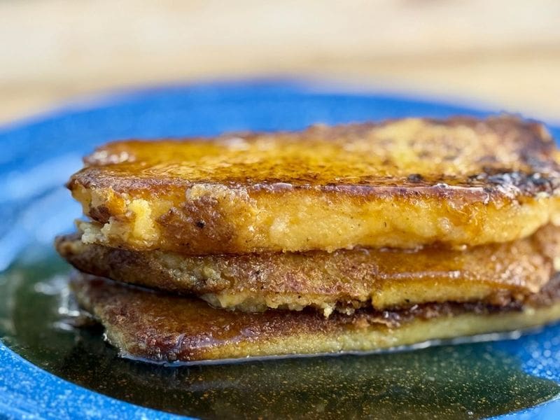 Golden-brown pan-fried cornmeal mush slices on a ceramic plate with butter melting on top, set on a wooden kitchen table in warm natural light