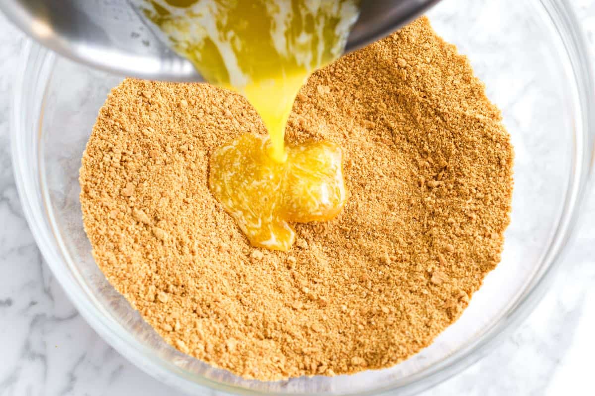 Graham cracker crumbs, melted butter, sugar, and a pinch of salt measured on a kitchen counter with a mixing bowl and spoon