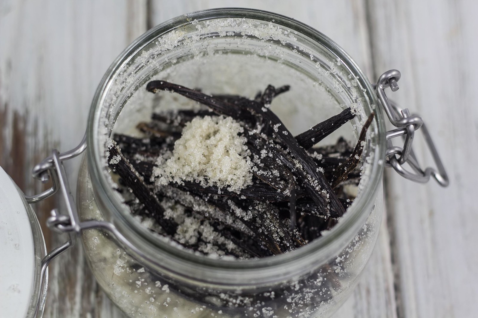 Granulated sugar in a bowl next to whole vanilla beans and a small glass jar on a clean kitchen counter