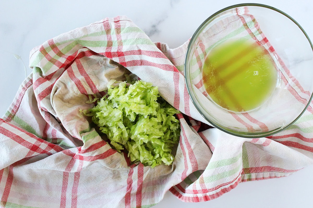 Grated cucumber wrapped in a clean kitchen towel being squeezed over a bowl on a bright kitchen counter