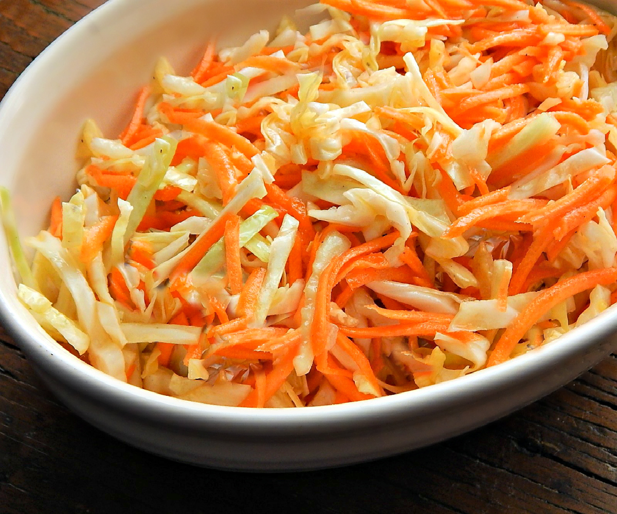 Green cabbage, a whole carrot, and a small onion on a clean kitchen counter with measuring cups and a mixing bowl ready for prep