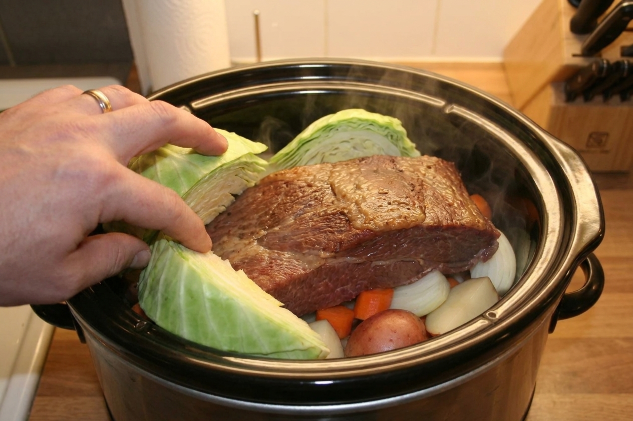 Green cabbage wedges being tucked into a slow cooker around a cooked corned beef brisket with steam rising