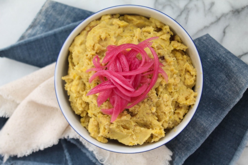 Green plantains, a red onion, a small bowl of vinegar, butter, and salt arranged on a bright kitchen counter