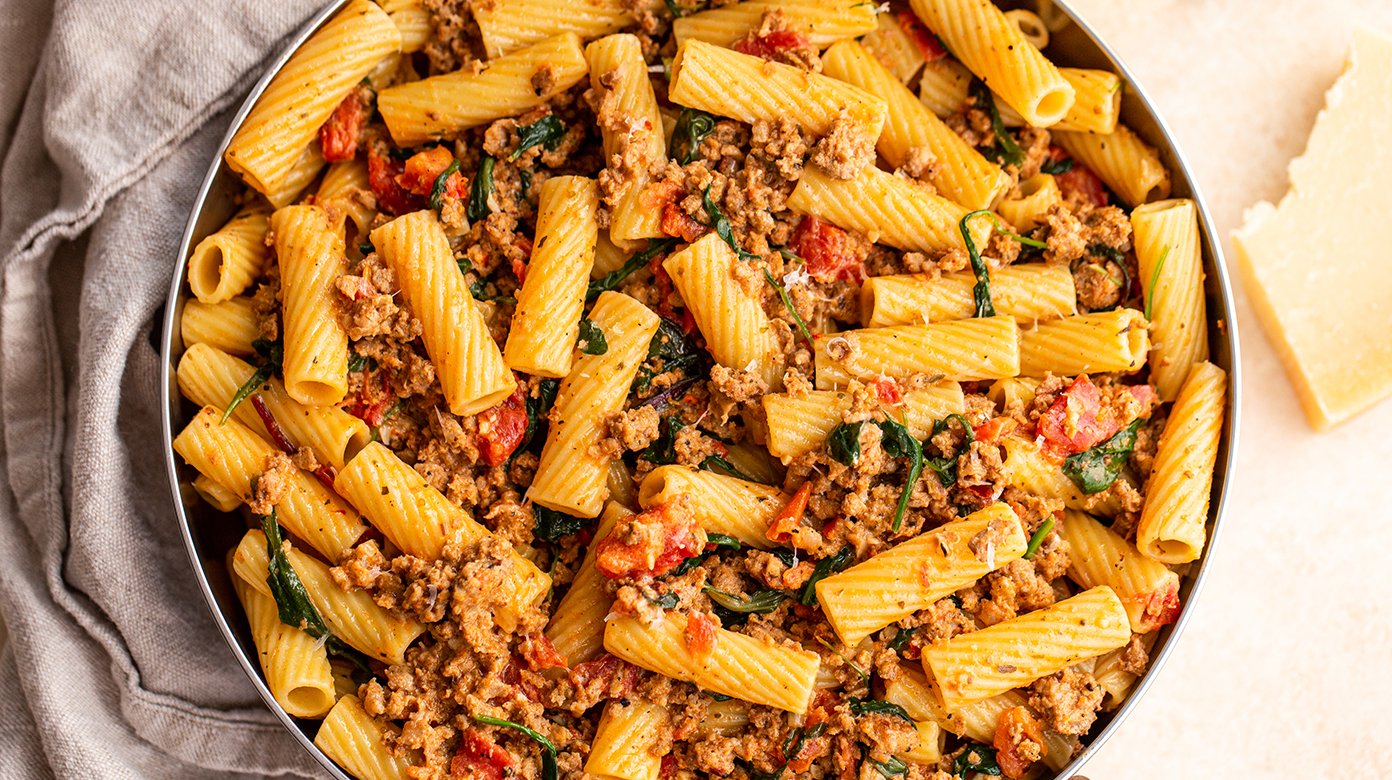 Ground Italian sausage, pasta, garlic, Parmesan, spinach, sun-dried tomatoes, and cream arranged on a clean kitchen counter with a cutting board and small bowls