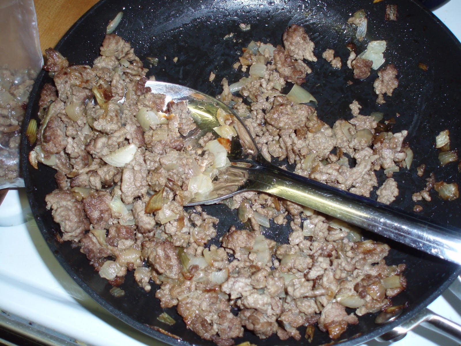 Ground beef and diced onion browning in a large skillet on a stovetop, with a wooden spoon stirring in a home kitchen
