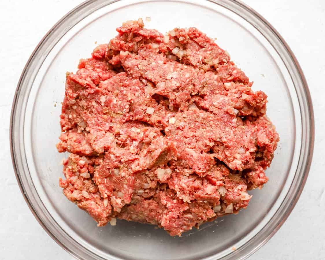 Ground beef, breadcrumbs, egg, grated onion, and seasonings combined in a large stainless steel bowl on a kitchen counter