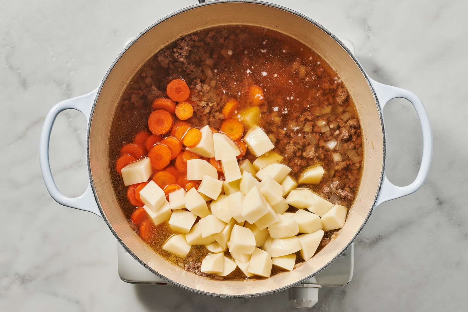 Ground beef browning in a large Dutch oven with diced onion and carrots on a stovetop, with a wooden spoon stirring