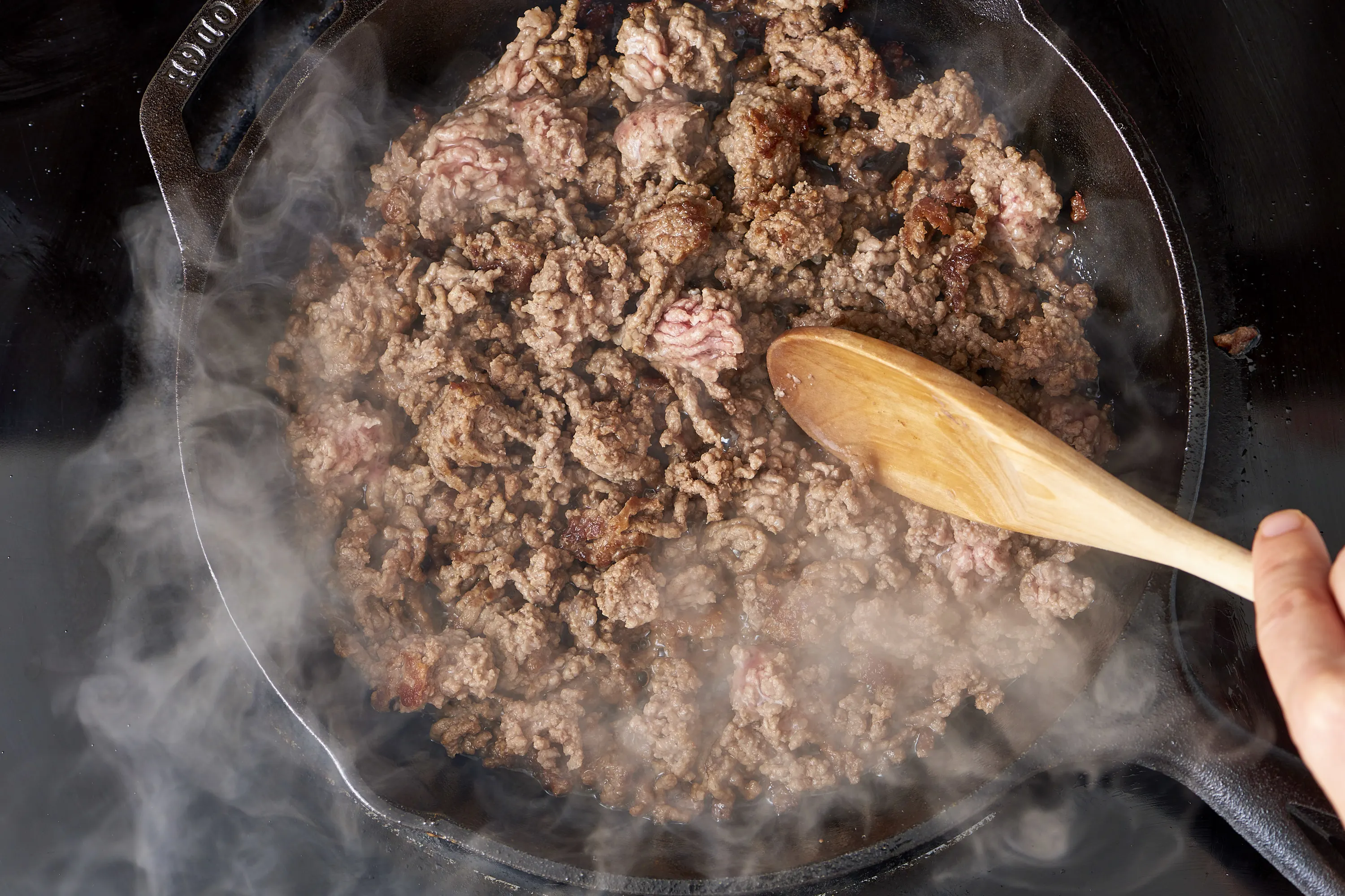 Ground beef browning in a large skillet on a stove with a wooden spoon stirring