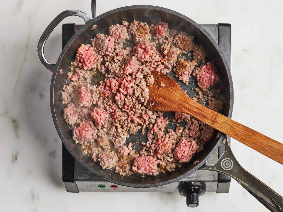 Ground beef browning in a large skillet on a stovetop as a wooden spoon breaks it into small crumbles