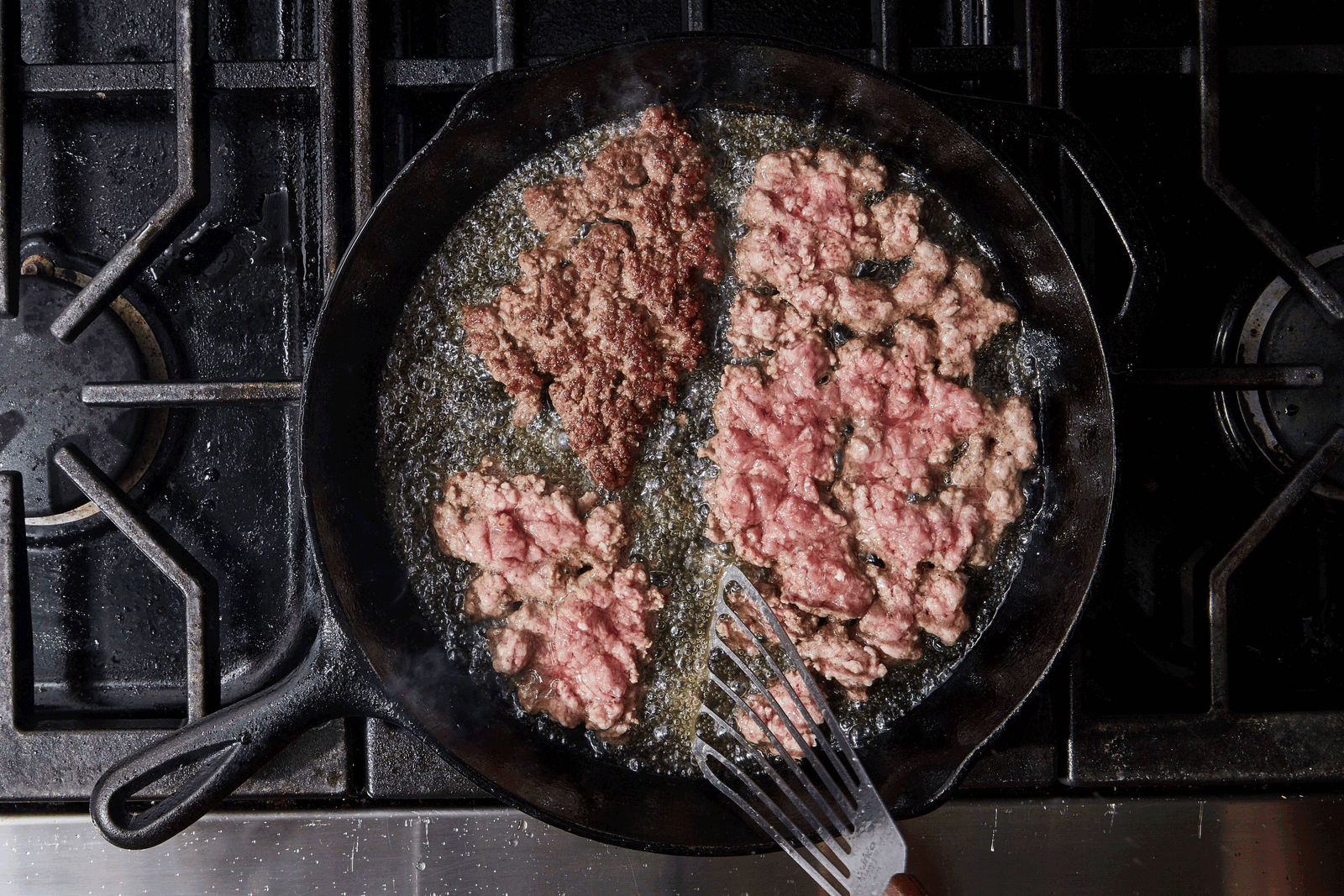 Ground beef browning in a wide skillet on a stovetop with a wooden spoon nearby, showing crisp browned bits forming