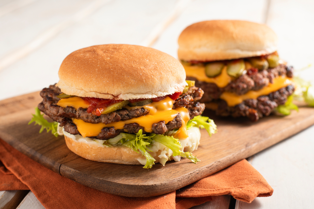 Ground beef, burger buns, sliced American cheese, onions, pickles, mayo, ketchup, mustard, and spices arranged on a kitchen counter with a cutting board