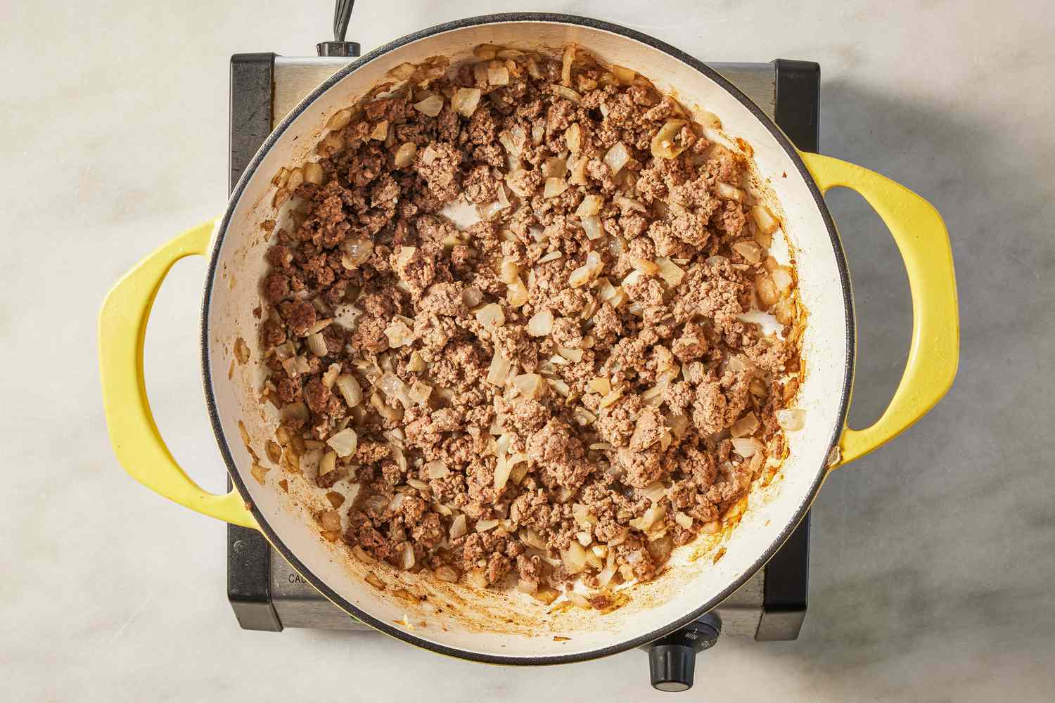 Ground beef, chopped onion, minced garlic, and small bowls of spices arranged on a kitchen counter with a cast iron skillet in the background