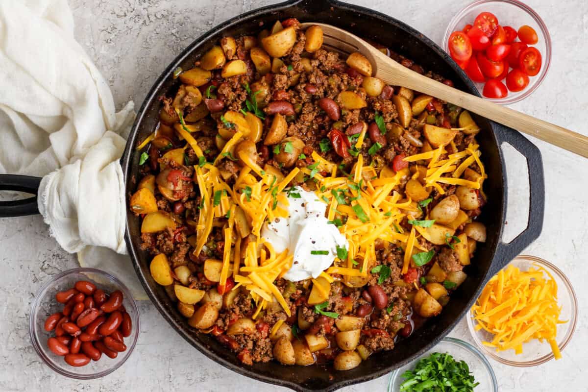 Ground beef, diced onion, garlic, spices, beans, and canned tomatoes staged near a skillet on a stove in a bright home kitchen