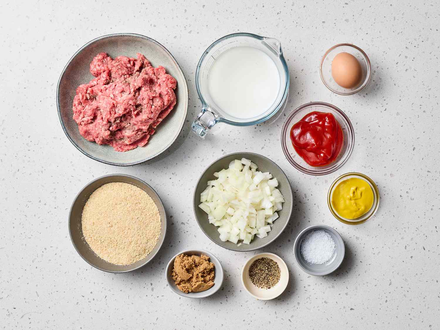 Ground beef, ground pork, eggs, breadcrumbs, diced onion, milk, seasonings, and BBQ sauce arranged on a clean kitchen counter in a single scene