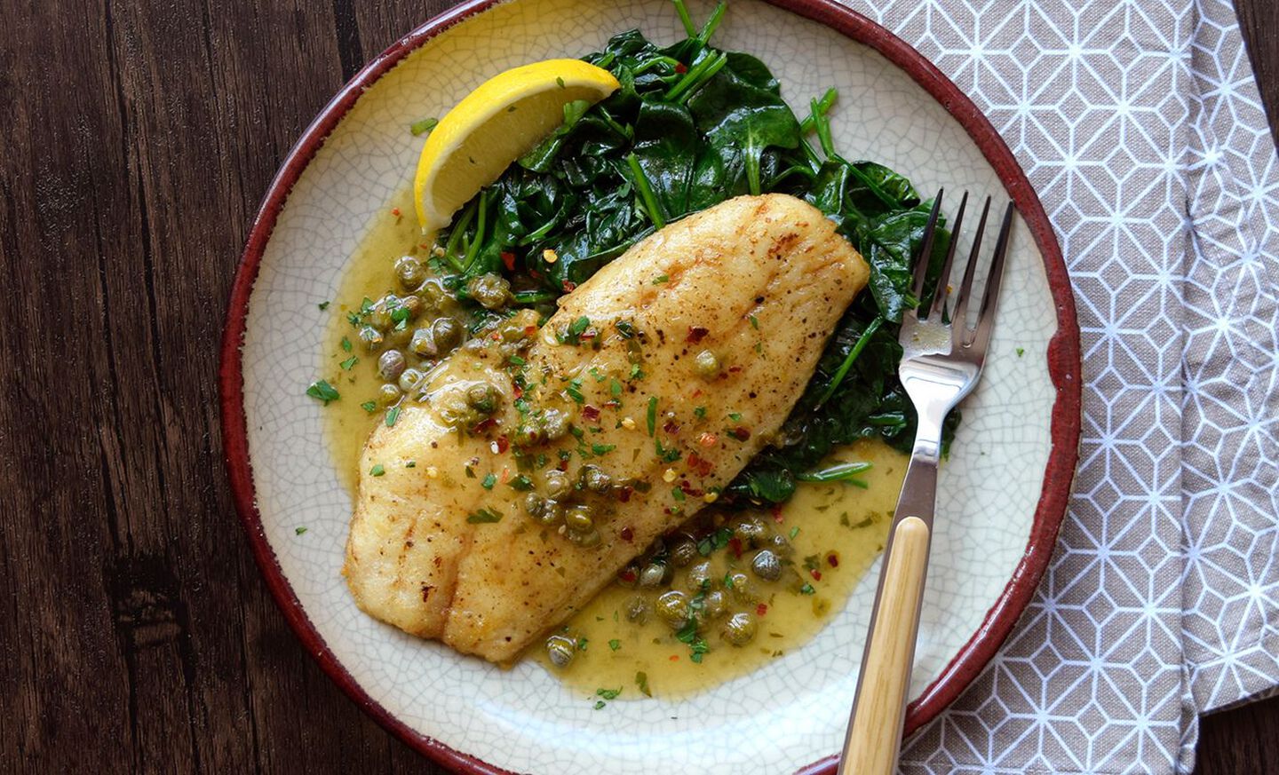 Grouper fillets patted dry and seasoned with salt and pepper on a plate next to a small bowl of flour and a lemon on a kitchen counter