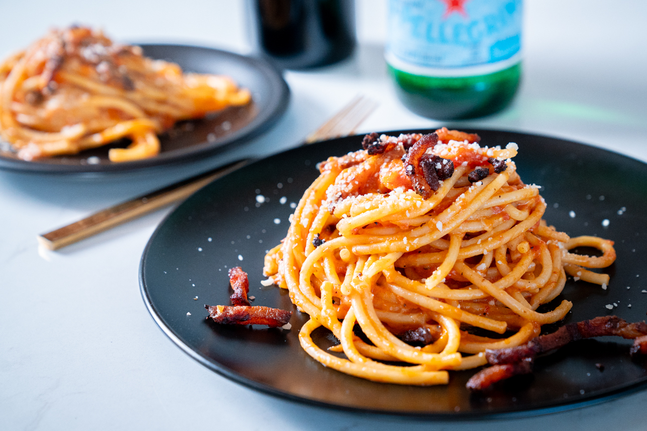 Guanciale, a can of whole peeled tomatoes, pecorino romano, dried bucatini, and a small bowl of red pepper flakes arranged on a clean kitchen counter in soft natural light