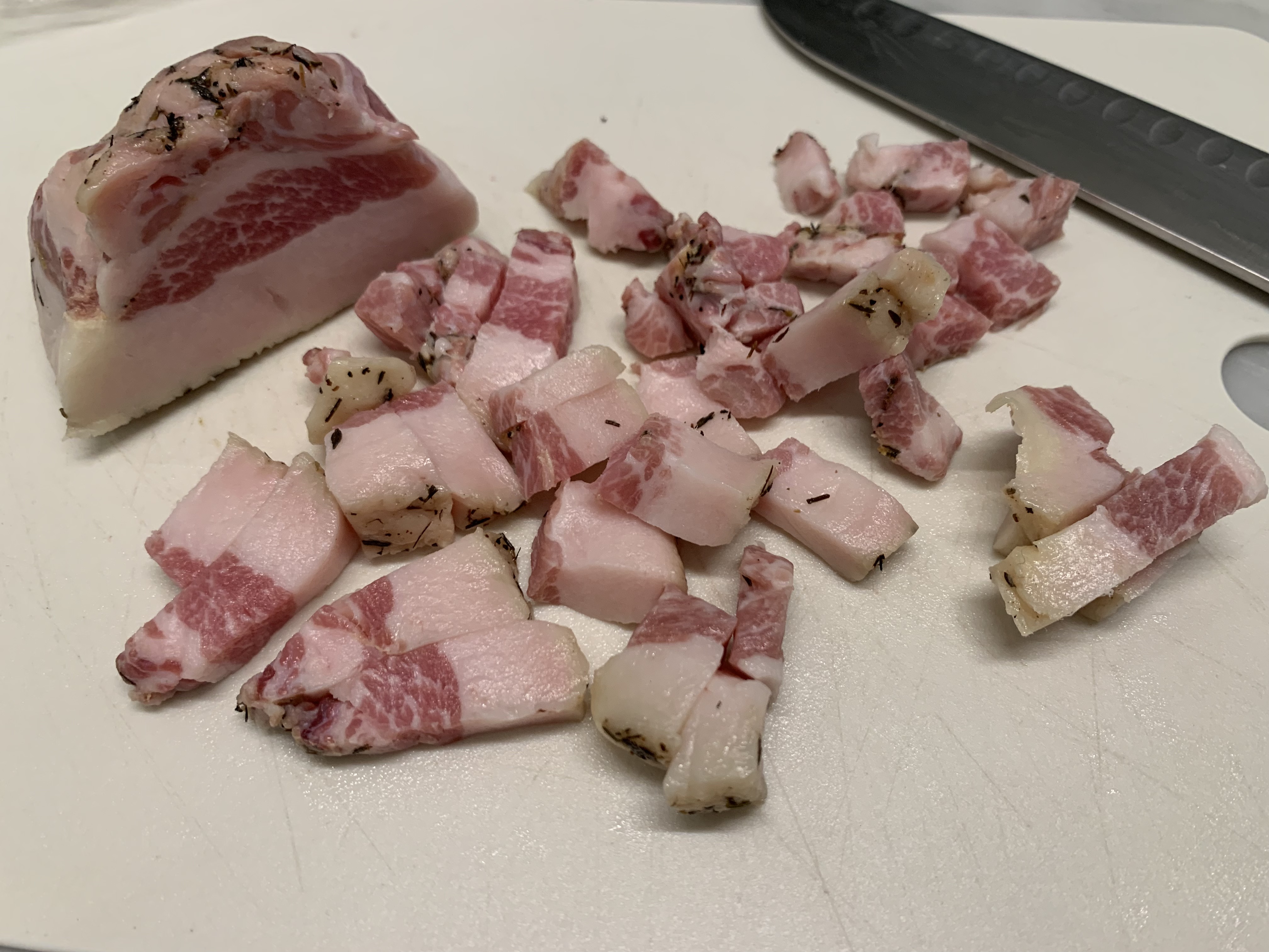 Guanciale cut into small batons on a wooden cutting board with a chef's knife, ready to render in a skillet