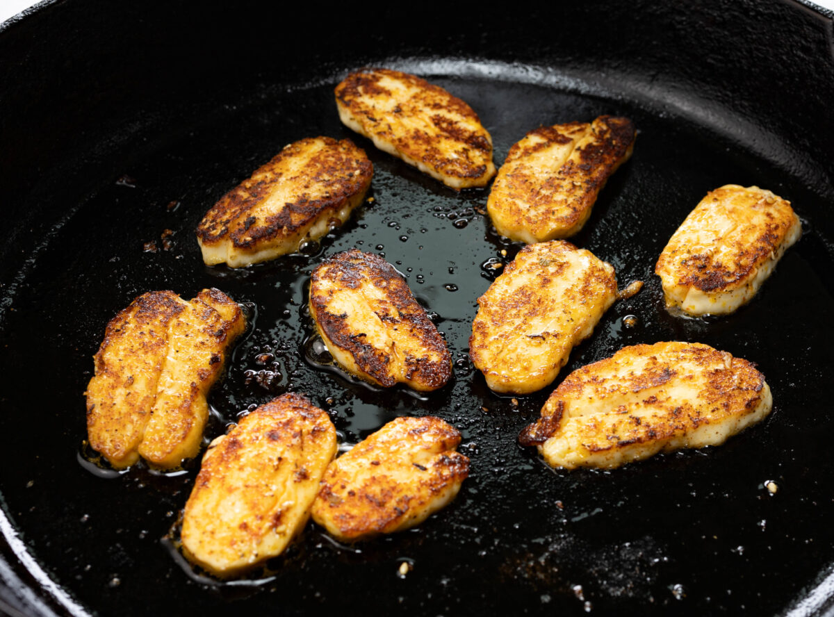 Halloumi slices browning in a cast iron skillet on a home stove with tongs resting nearby