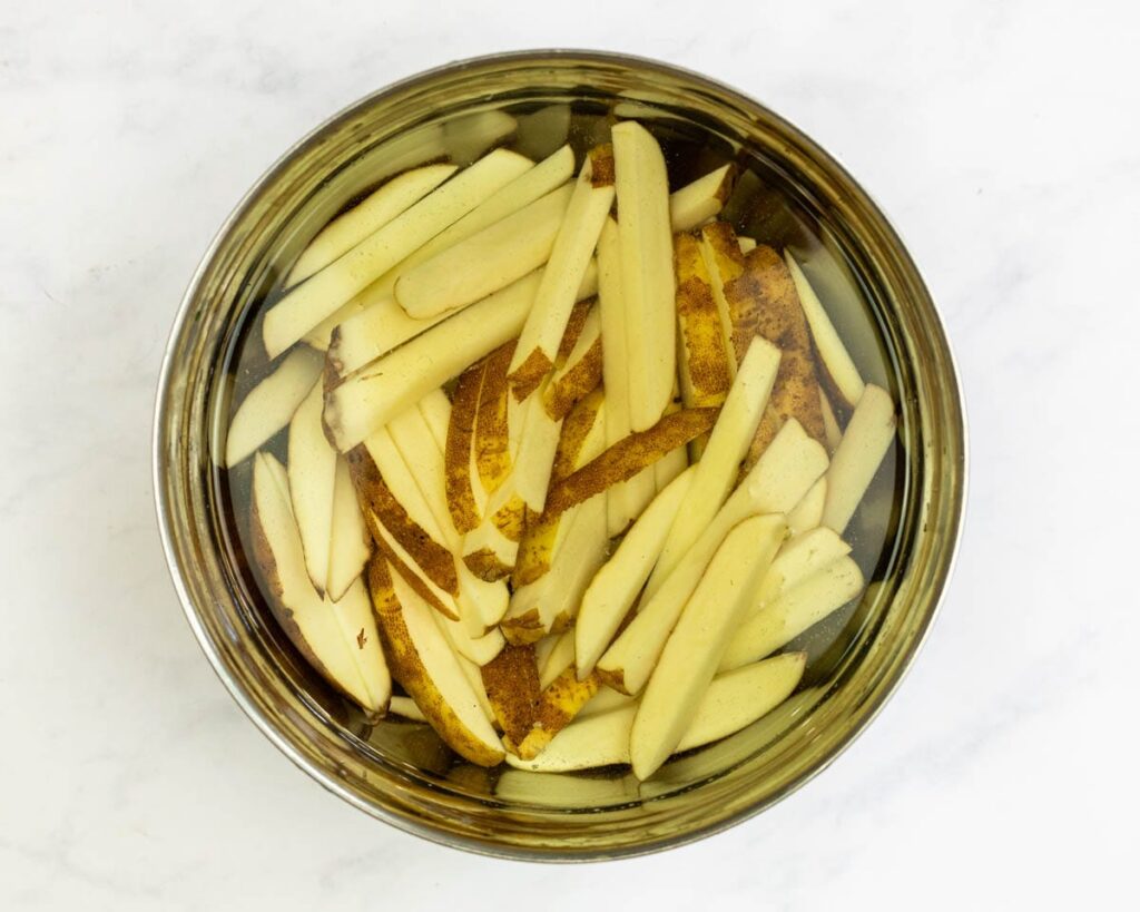 Hand-cut russet potato fries soaking in a clear bowl of cold water on a kitchen counter