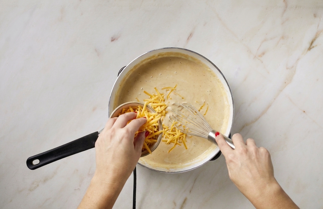 Hand sprinkling shredded cheddar into a saucepan of creamy sauce while stirring, cheese beginning to melt
