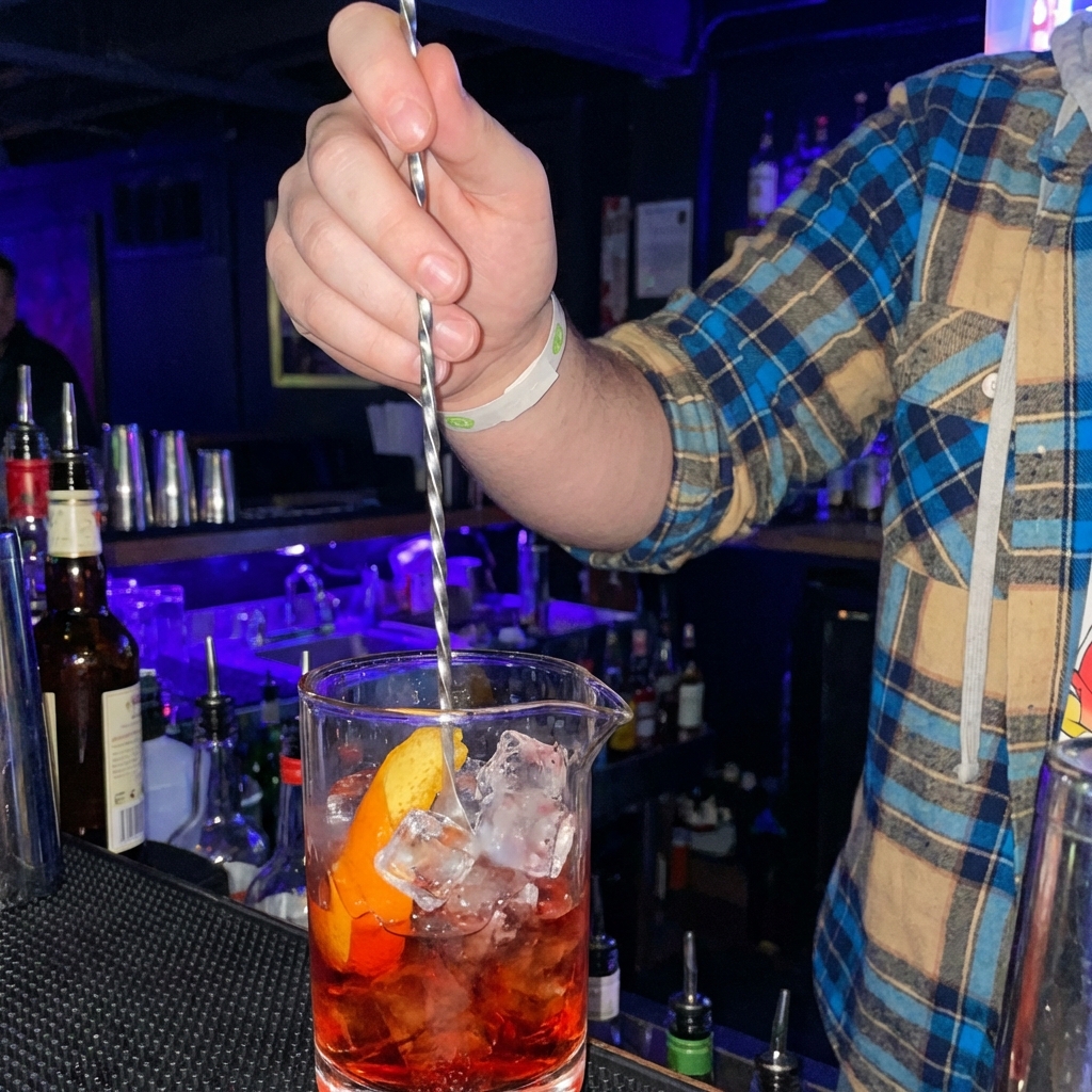 Hand stirring a Negroni-style drink in a mixing glass with ice and a long bar spoon