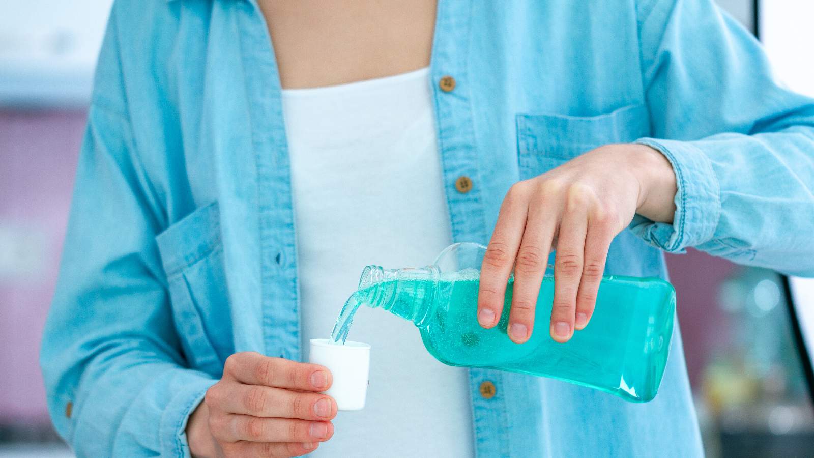 Hands at a kitchen counter measuring a dose of prescribed mouth rinse into a small medicine cup under warm natural light