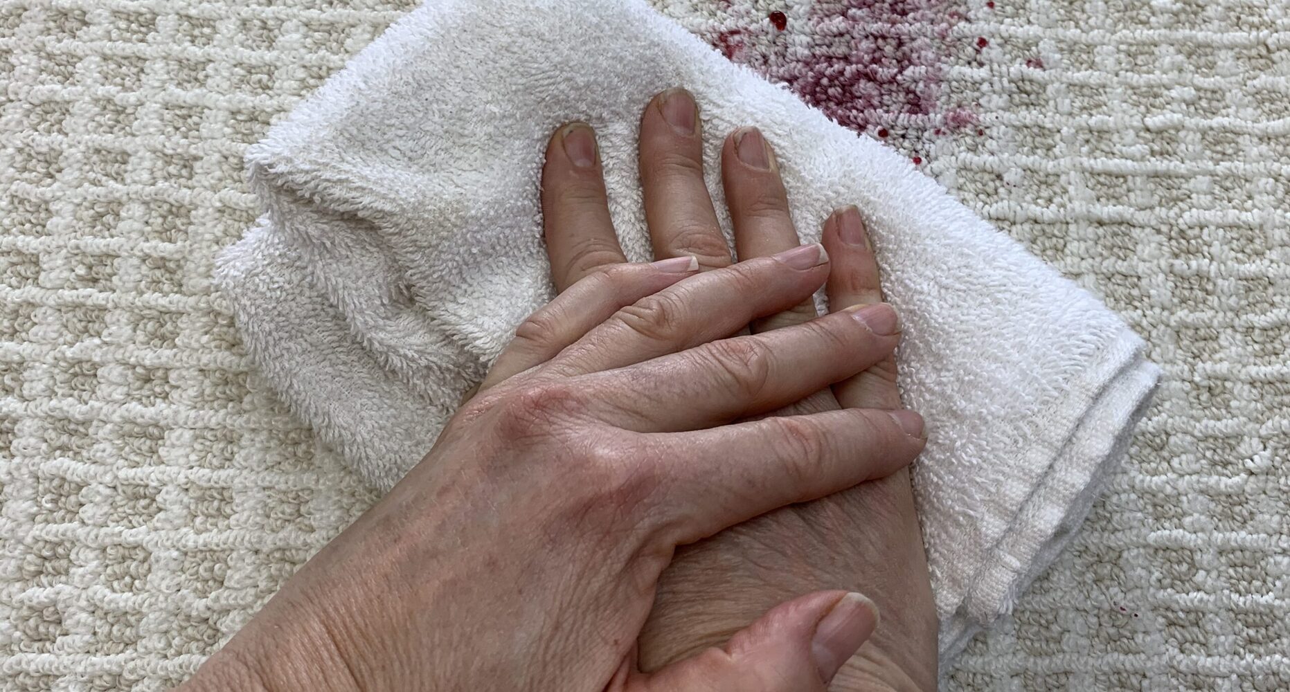 Hands blotting a fresh spill on beige carpet with a clean white towel, seen from above in natural light