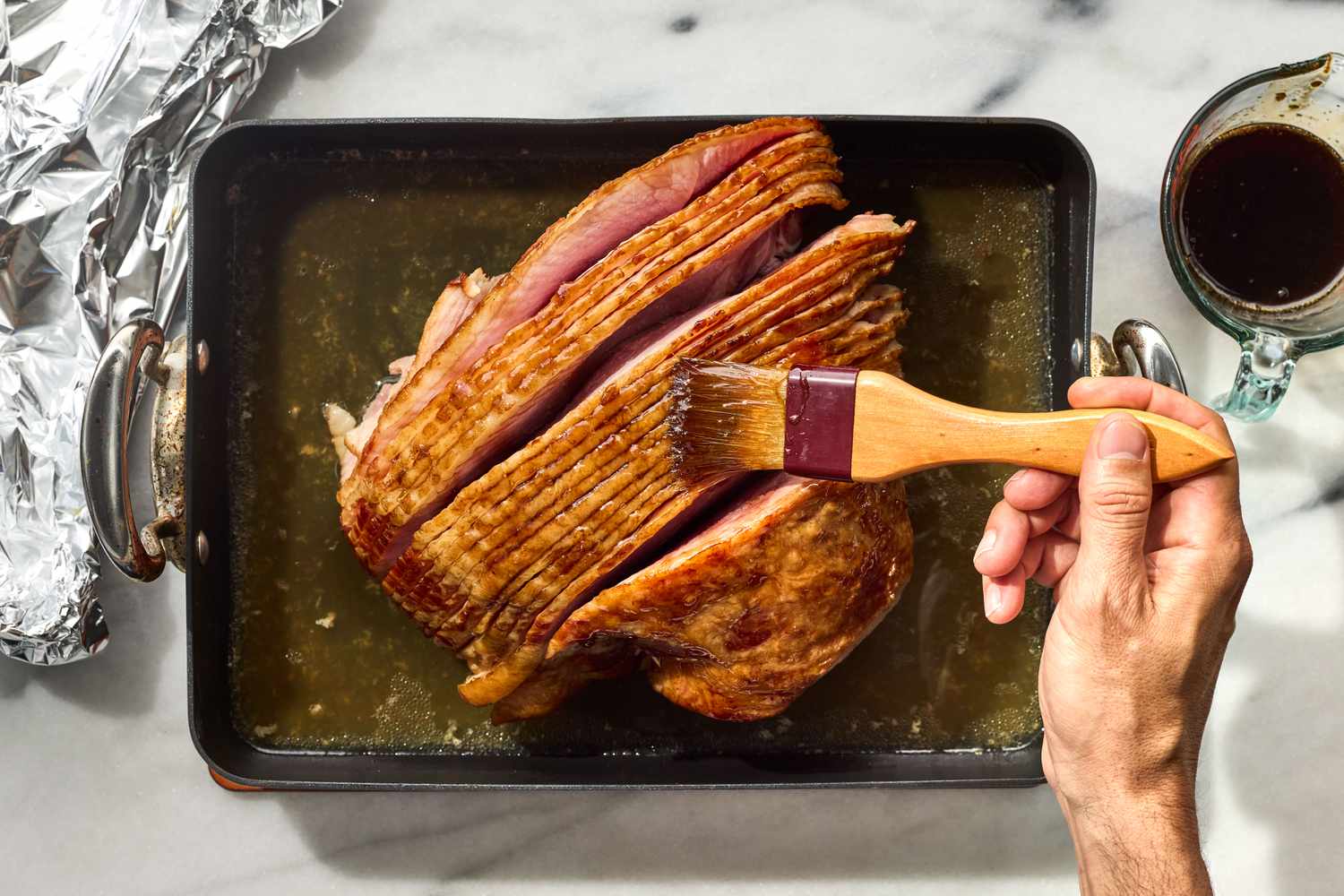 Hands brushing shiny brown sugar glaze over a warm spiral ham in a roasting pan on a kitchen counter, with foil loosely pulled back