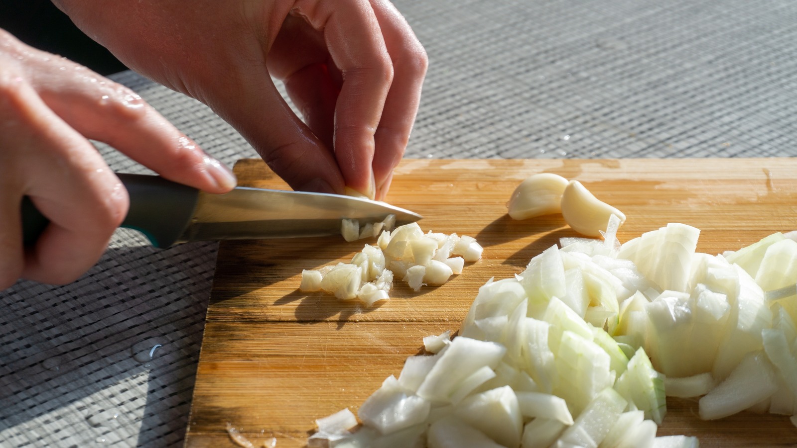 Hands chopping a yellow onion and mincing garlic on a wooden cutting board on a home kitchen counter