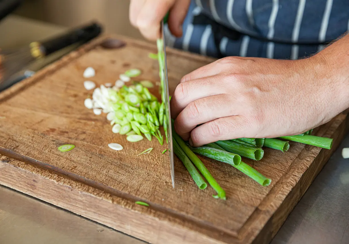 Hands chopping scallions and onion on a wooden cutting board with thyme and garlic nearby on a kitchen counter