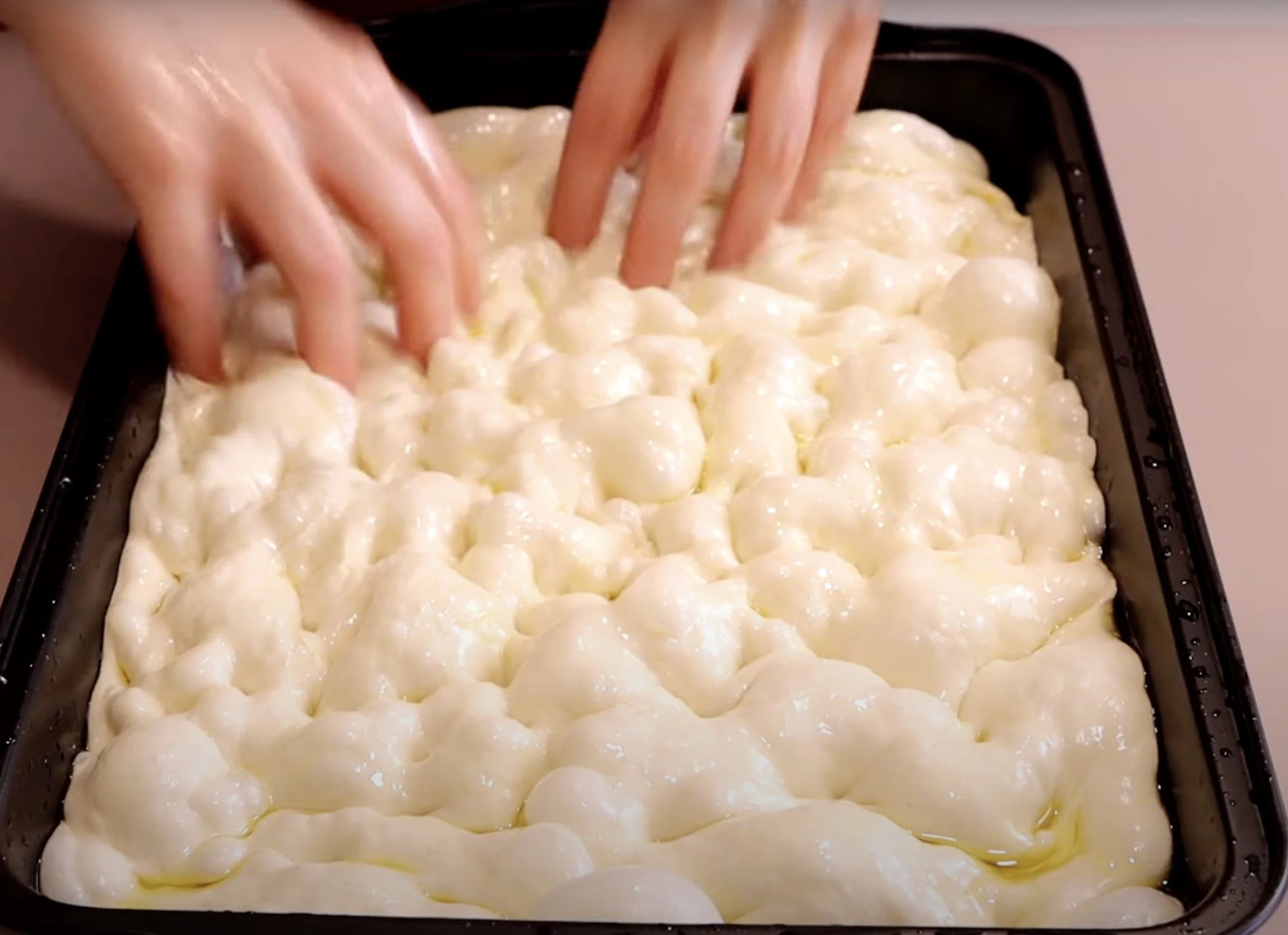 Hands dimpling focaccia dough in an oiled pan